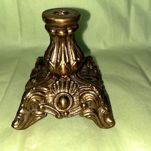 May include: Ornate brass candlestick holder with a detailed, scrolled base and a fluted column. The antique-style candle holder has a round top for holding a candle. The brass finish gives it a vintage look.