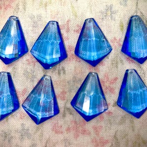 Puede incluir: Ocho cuentas de vidrio azul facetadas en forma de lágrima. Cada cuenta tiene una base puntiaguda y un pequeño agujero para ensartar. Las cuentas están dispuestas sobre una tela estampada floral.