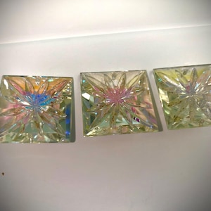 May include: Three square, iridescent glass coasters. Each coaster features a starburst design with a central flower-like pattern. The glass reflects a spectrum of colors, including pink, blue, and yellow, creating a shimmering effect.