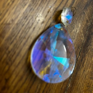 May include: A clear, teardrop-shaped crystal pendant with iridescent blue and purple hues. The pendant has a small, faceted crystal at the top with a metal loop. The crystal is resting on a wooden surface.