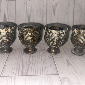 May include: A set of four small, chalice-shaped egg cups featuring a dark, weathered metallic finish with raised leaf and berry detailing. Each cup has a wide, textured bowl, a narrow stem, and a circular base, showcasing a distressed patina.