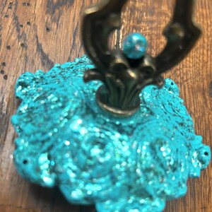 May include: A decorative teal-colored object with a textured, glittery surface. The object has an ornate, dark bronze-colored metal handle and a small, teal-colored bead at the top. The base has a scalloped edge.