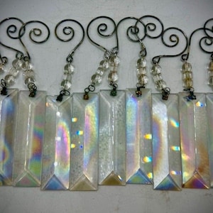 May include: A collection of rectangular, clear crystal prisms with iridescent color reflections. Each prism hangs from a decorative metal hook with a spiral design and a strand of clear beads. The prisms are arranged in a row.