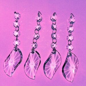 May include: Four clear crystal chandelier pendants with a leaf-like shape and a series of faceted beads. The pendants are suspended from metal hooks and chains, set against a soft purple background. The crystals reflect light, creating a sparkling effect.
