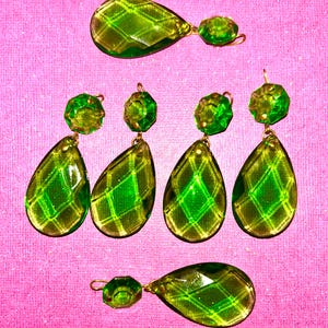 Könnte beinhalten: Mehrere grüne und gelbe Kristallohrringe mit Rautenmuster vor rosa Hintergrund. Die Ohrringe haben eine Tropfenform und sind mit einem kleineren, facettierten Kristall verbunden. Modeschmuck.