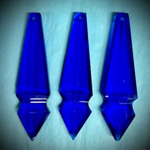 May include: Three vibrant blue glass pendants with a pointed, elongated shape. Each pendant has a small hole at the top and a diamond-shaped base. The pendants are displayed against a light blue background.