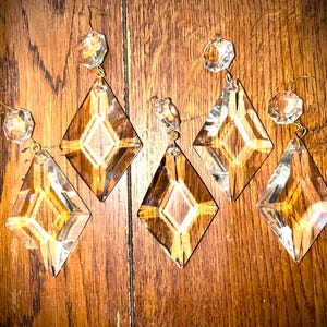 Może przedstawiać: Pięć przezroczystych kryształowych pryzmatów do żyrandola o kształcie rombu i fasetowanych okrągłych akcentach. Pryzmaty zawieszone są na złotych drucianych haczykach. Kryształy są prezentowane na tle ciemnego drewna.