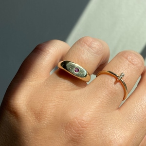 May include: Two gold rings on a hand. One ring is a thick band with a small, round, pink gemstone. The other ring is a thin band with a small, clear gemstone. The rings are displayed in natural sunlight.