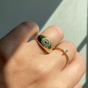 Puede incluir: Un anillo de oro con una piedra preciosa roja engastada en el centro. El anillo se lleva en un dedo con una fina banda dorada en el dedo anular.