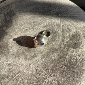 May include: A small, silver-colored pendant with a rounded shape and a loop for a chain. The pendant features two small, dark blue gemstones. The pendant is resting on a gray background with a detailed, circular pattern.