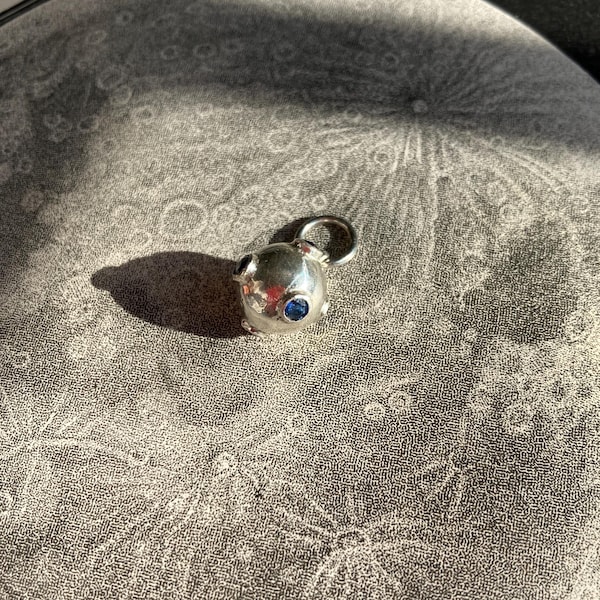 May include: A small, spherical silver charm featuring a single, round, deep blue gemstone set into its polished surface. The charm includes a small circular jump ring at the top for attachment, reflecting light with a bright, metallic sheen.