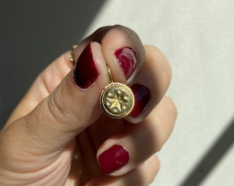 May include: A gold-colored earring with a starburst design. The earring has a circular shape with a textured border. The starburst is in the center of the earring. The background is a light color with shadows.