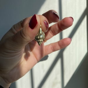 May include: A small, gold-colored seashell pendant. The pendant is held up against a white background with shadows. The seashell has a detailed, textured surface and a loop for a chain. The pendant is a piece of jewelry.