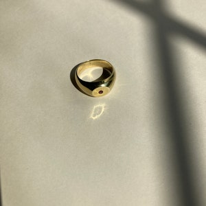 May include: A gold ring with a red gemstone set in the center. The ring is a simple, classic design.