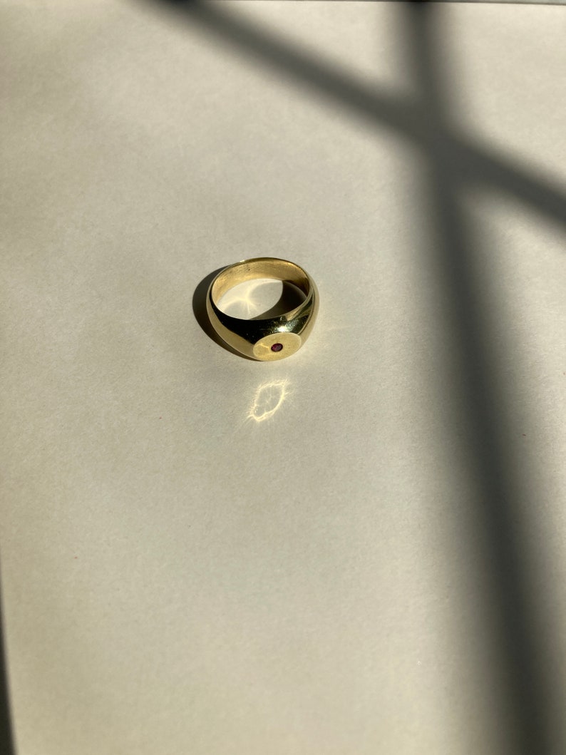 May include: A gold ring with a red gemstone set in the center. The ring is a simple, classic design.
