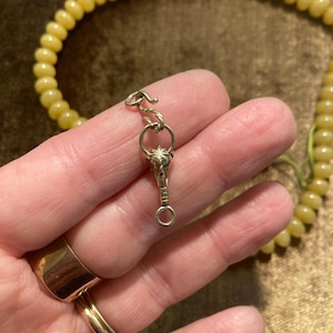 May include: A gold-colored necklace clasp with a detailed pendant featuring a starburst design. The clasp is connected to a strand of yellow beads. The image shows a close-up of the clasp and a portion of the necklace.