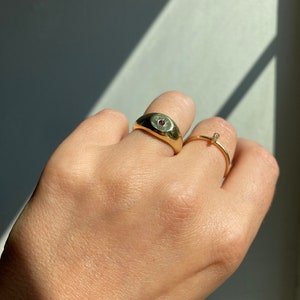 May include: A gold ring with a single red gemstone set in the center of the band. The ring is worn on the index finger of a hand.