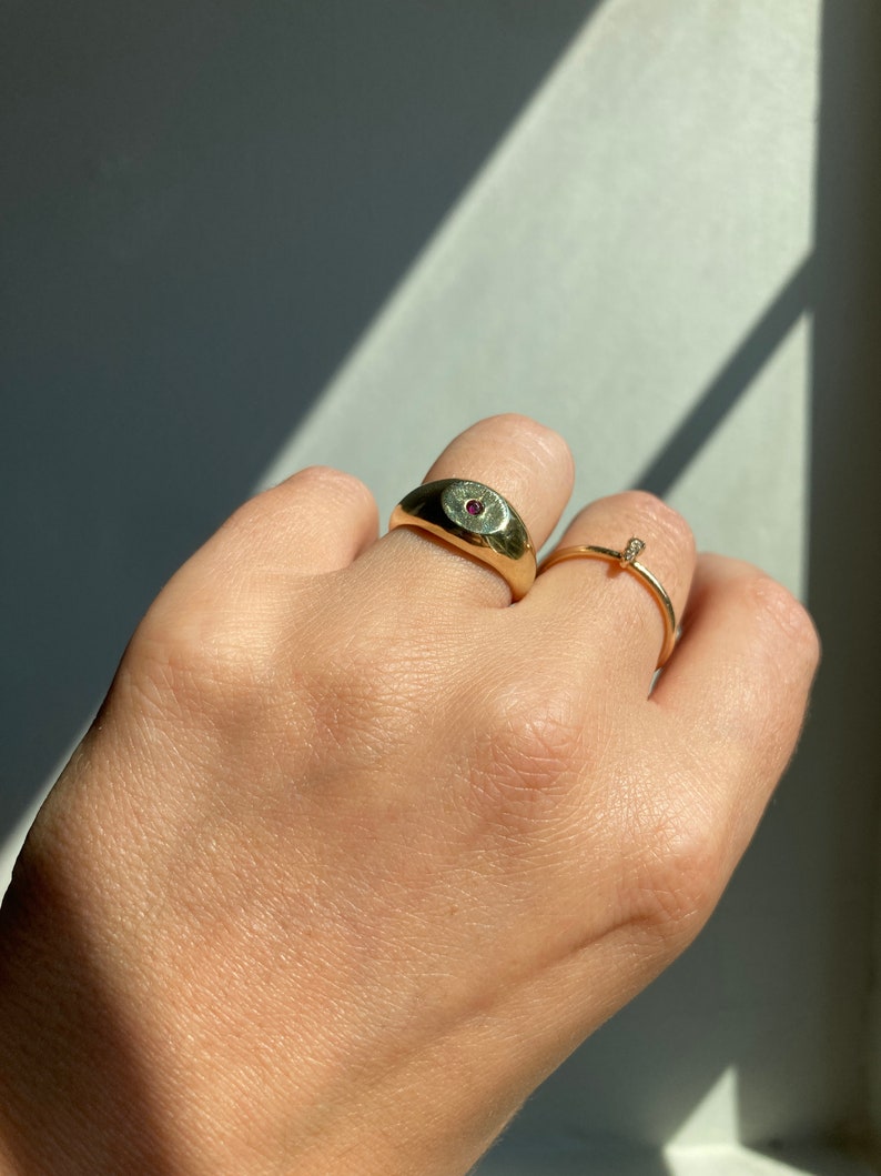 May include: A gold ring with a single red gemstone set in the center of the band. The ring is worn on the index finger of a hand.