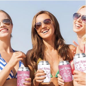 May include: A group of smiling women wearing sunglasses hold beverage insulators. The insulators are white and pink, with the text "Last Rodeo" and a graphic of a cowboy on them. The background is a sunny beach scene.
