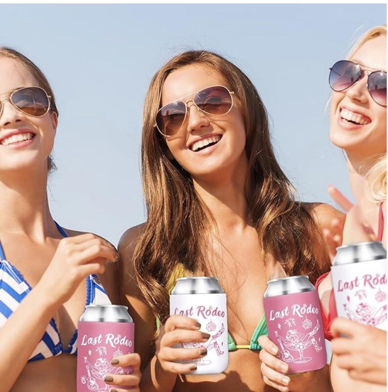 May include: A group of smiling women wearing sunglasses hold beverage insulators. The insulators are white and pink, with the text "Last Rodeo" and a graphic of a cowboy on them. The background is a sunny beach scene.