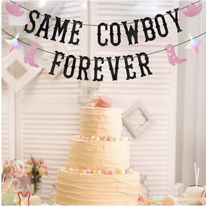 Può includere: Una torta a tre strati con decorazioni rosa e bianche è posizionata sotto uno striscione con la scritta "SAME COWBOY FOREVER". Lo striscione presenta lettere nere glitterate, stivali da cowboy rosa e stelle su sfondo bianco.