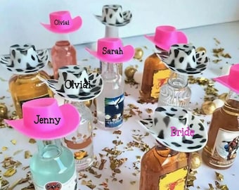 May include: Miniature liquor bottles topped with pink cowboy hats and black and white cow print hats. Some hats have names like "Olviai", "Sarah", "Jenny", and "Bride". Gold confetti is scattered around the bottles.