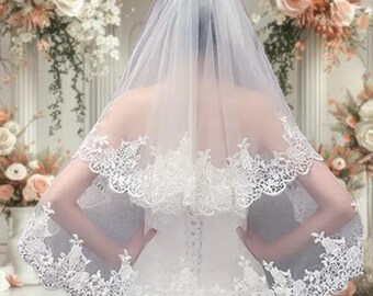 May include: A white, two-tiered bridal veil with delicate lace trim. The veil is sheer, allowing a glimpse of the wearer's back and dress. The lace features an intricate floral design, adding a touch of elegance to the bridal accessory.