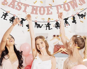 May include: A party banner with the text "LAST HOE DOWN" in glittery rose gold letters. Black silhouette cutouts of figures in various poses hang below. Confetti and three women celebrating with champagne glasses.