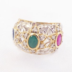 May include: A gold ring featuring a delicate filigree design with three oval gemstones: a green emerald, a pink ruby, and a blue sapphire.