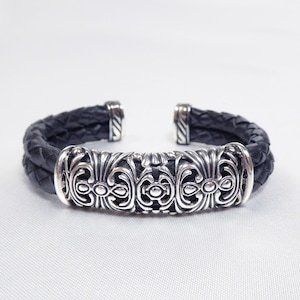 May include: A black leather bracelet with a silver ornate design. The bracelet is braided and has a silver clasp.