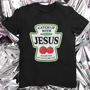 May include: Black t-shirt with a ketchup bottle design that says "Catch Up With Jesus" and "Psalm 37:4". The bottle has two red tomatoes on it and the text "Blessed From My Head To-Ma-Toes".
