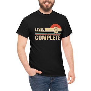 May include: A black t-shirt with a vintage-style graphic that says "Level 8 Complete" with a red, orange, and yellow sunset design and a white 8 ball.