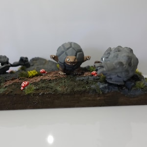 Puede incluir: Un diorama en miniatura con tres figuras grises con forma de piedra sobre una pequeña base terrosa. La escena incluye pequeñas setas rojas y blancas, vegetación verde y amarilla, y una criatura parecida a una tortuga.
