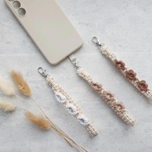Könnte beinhalten: Drei Makramee-Handy-Schlaufen mit Blumenakzenten in Creme- und Brauntönen. Jeder Riemen hat einen silberfarbenen Verschluss. Auf dem Bild ist auch eine beige Handyhülle zu sehen, zusammen mit getrocknetem Ziergras.