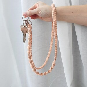 Könnte beinhalten: Ein pfirsichfarbenes Makramee-Schlüsselanhänger-Armband mit einem Schlüsselring, der mehrere Schlüssel hält. Das Armband besteht aus geknotetem Kordel und wird von einer Hand vor einem weißen Vorhanghintergrund gehalten.