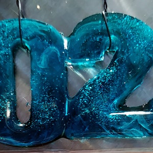 May include: A teal blue resin ornament shaped like the numbers 2023. The ornament has a swirled, marbled effect and is decorated with glitter.