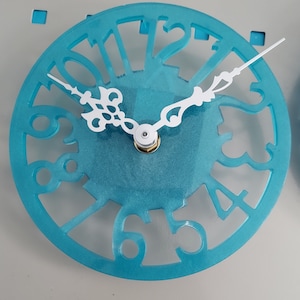 May include: A turquoise blue clock face with white Roman numeral hour markers and a white clock hand. The clock face is made of acrylic and has a glittery finish.