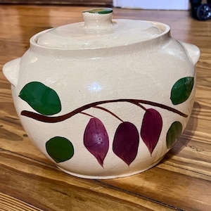 May include: A cream-colored ceramic casserole dish with a matching lid, featuring a hand-painted brown branch motif with green leaves and purple fruit. The round pot has two side handles and a visible crackle glaze finish across the surface.