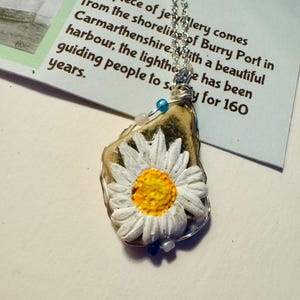 Puede incluir: Una margarita blanca con un centro amarillo está encerrada en un colgante de resina transparente. El colgante está unido a una cadena de plata. El collar está sobre una tarjeta blanca con texto sobre el origen de la pieza.