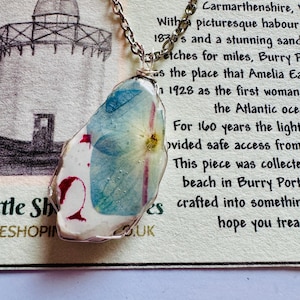 Può includere: Collana con catena d'argento e ciondolo in resina. Il ciondolo contiene fiori secchi blu e gialli, dettagli rossi su base bianca. Lo sfondo include testo su Burry Port, Carmarthenshire.