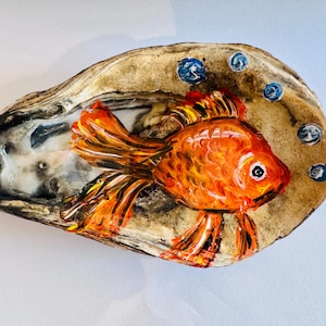 Könnte beinhalten: Handbemalte Austernschalenkunst mit einem orangefarbenen Goldfisch mit detaillierten Flossen und Schwanz. Der Fisch ist vor einem beigen Hintergrund mit blauen Blasenakzenten platziert. Die Schale hat ein natürliches, strukturiertes Aussehen und ist ein Unikat.