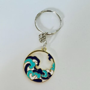 May include: A gold-coloured key chain with a round charm featuring a blue and white wave design. The charm is attached to a silver key ring.