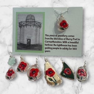 Può includere: Un set di sei ciondoli per gioielli fatti a mano con papaveri rossi dipinti su vetro di mare. I ciondoli sono attaccati a un filo d'argento e sono pronti per essere infilati su collane o braccialetti. L'immagine include anche una carta con un disegno di un faro e un testo sull'origine del vetro di mare.