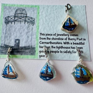 May include: Three silver charm necklaces with blue and white glass pendants depicting sailboats. The charms are displayed on a white card with a hand-drawn image of a lighthouse and the text "This piece of jewellery comes from the shoreline of Burry Port in Carmarthenshire. With a beautiful harbour, the lighthouse has been guiding people to safety for 100 years."