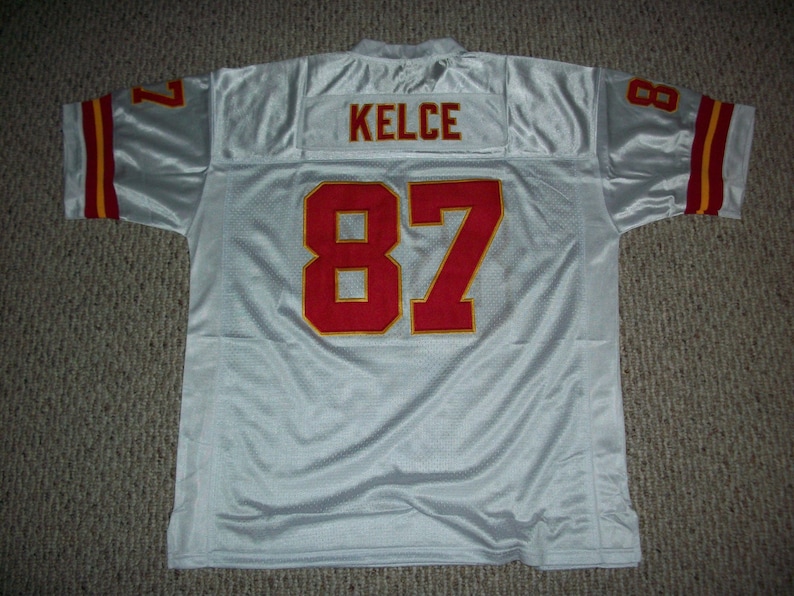 May include: White football jersey with red and gold trim. The back of the jersey has the number 87 and the name "KELCE" in red.