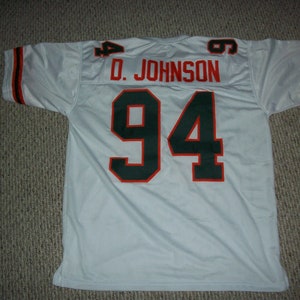 K&ouml;nnte beinhalten: Wei&szlig;es Football-Trikot mit gr&uuml;nen und orangen Verzierungen. Auf der R&uuml;ckseite des Trikots befindet sich die Nummer "94" in Gr&uuml;n mit orangenen Umrandungen. Der Name "D. JOHNSON" ist &uuml;ber der Nummer aufgedruckt.