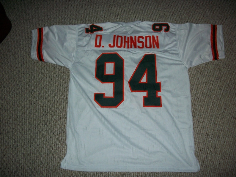 K&ouml;nnte beinhalten: Wei&szlig;es Football-Trikot mit gr&uuml;nen und orangen Verzierungen. Auf der R&uuml;ckseite des Trikots befindet sich die Nummer "94" in Gr&uuml;n mit orangenen Umrandungen. Der Name "D. JOHNSON" ist &uuml;ber der Nummer aufgedruckt.