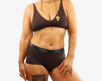 May include: A brown bra and matching boy shorts set. The shorts have a black waistband with the text "Y.O.U." in white. The bra has a v-neck design. The model is wearing jewelry and has a tiger tattoo on their thigh.
