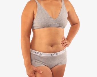 May include: A light gray bra and matching boy shorts set. The bra has a V-neck design. The boy shorts have a white waistband with the text "Y.O.U." printed on it. The set is made of a soft, comfortable material.
