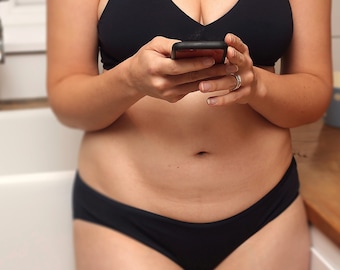 May include: A person wearing black underwear and a black bra, holding a black smartphone.